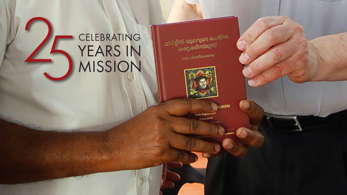 Two men hold Catechism, text reads "Celebrating 25 Years in Mission"